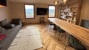 A cozy living room with a sofa, a coffee table, a wooden dining table, and chairs. There are two windows with a view of the countryside. A bookshelf filled with books and decorative items is against the wall. - Garibaldi 14 Suite (Chianciano Terme)