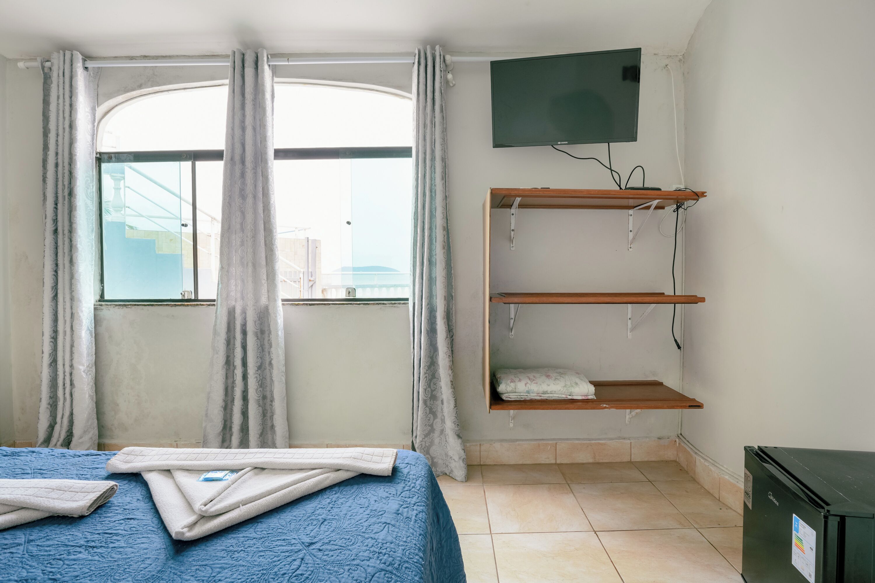 Une chambre à coucher avec un lit, un téléviseur fixé au mur, une fenêtre avec des rideaux et une petite étagère.