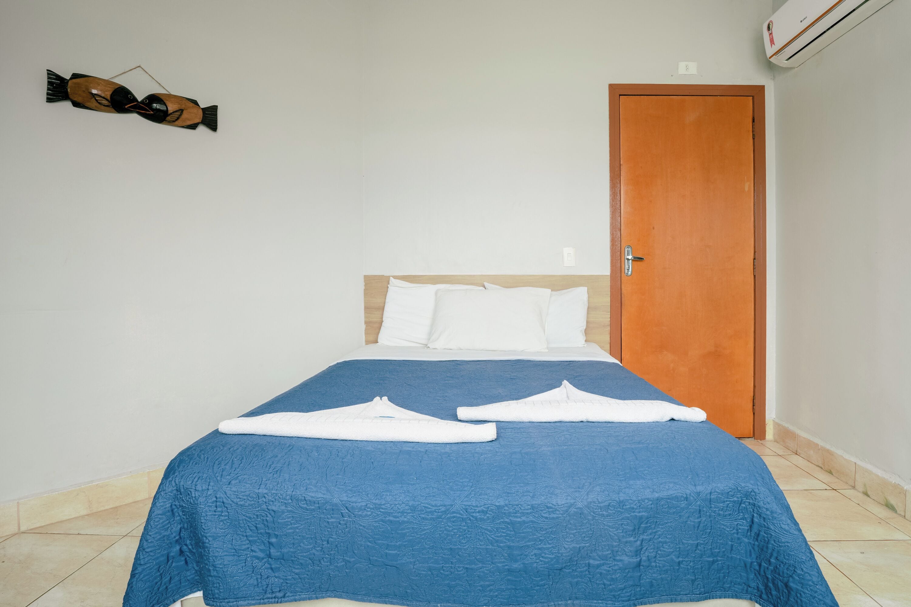 Una cama bien tendida con una colcha azul y almohadas blancas, una puerta de madera y un equipo de aire acondicionado instalado en la pared.