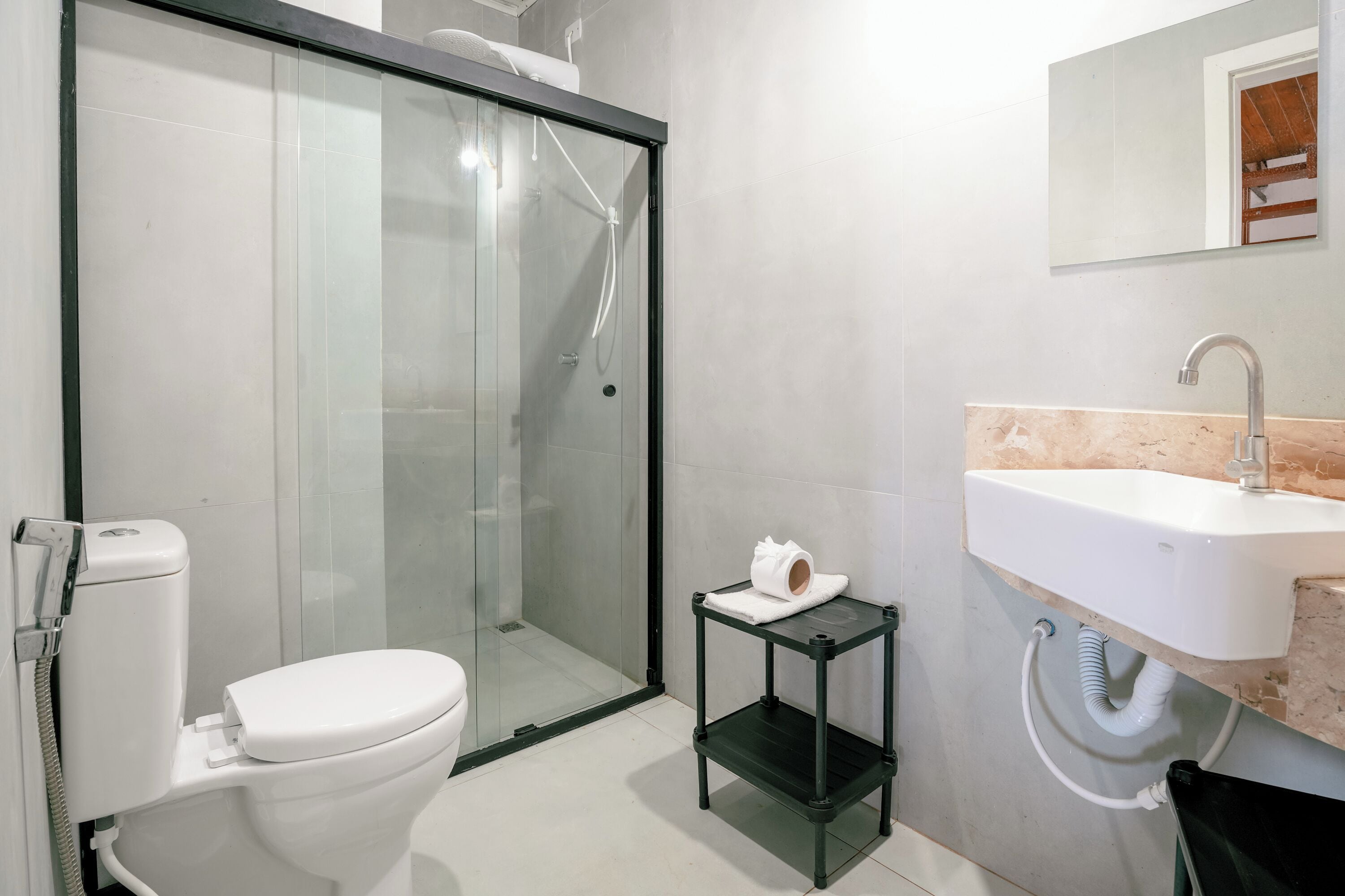 A modern bathroom with a glass shower enclosure, a toilet, a sink with a marble countertop, and a small black shelf.