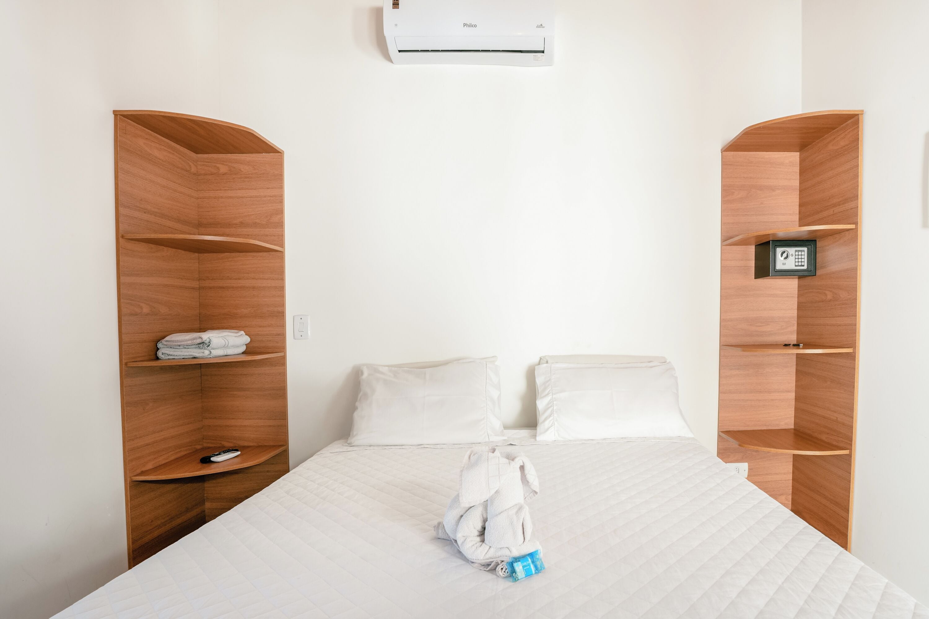 A hotel room with a bed, two pillows, a folded towel, a remote control, and a small shelf with a lockbox.