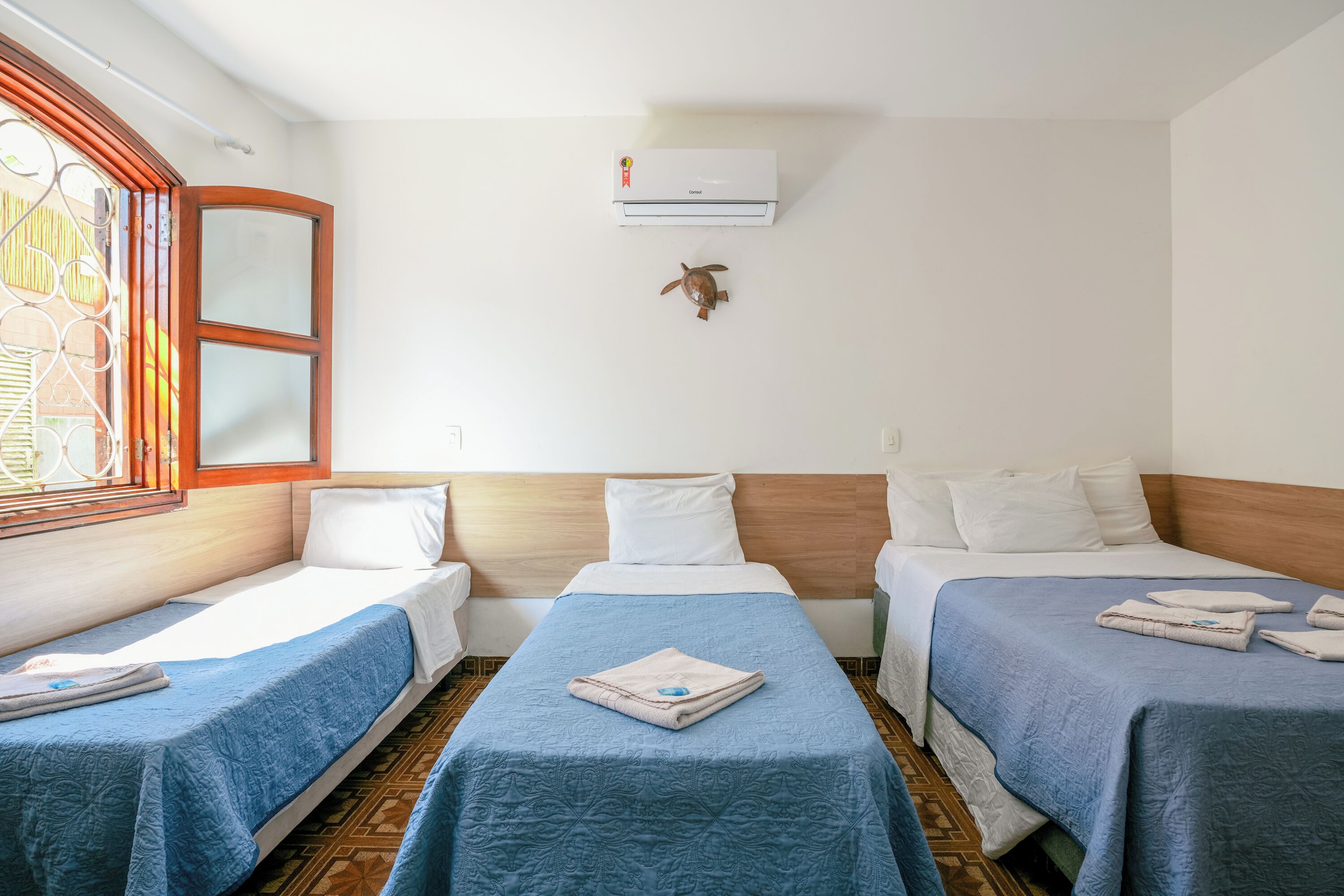 Una habitación de hotel con dos camas, un ventanal con celosía de hierro decorativa, un equipo de aire acondicionado y una decoración de tortuga en la pared.