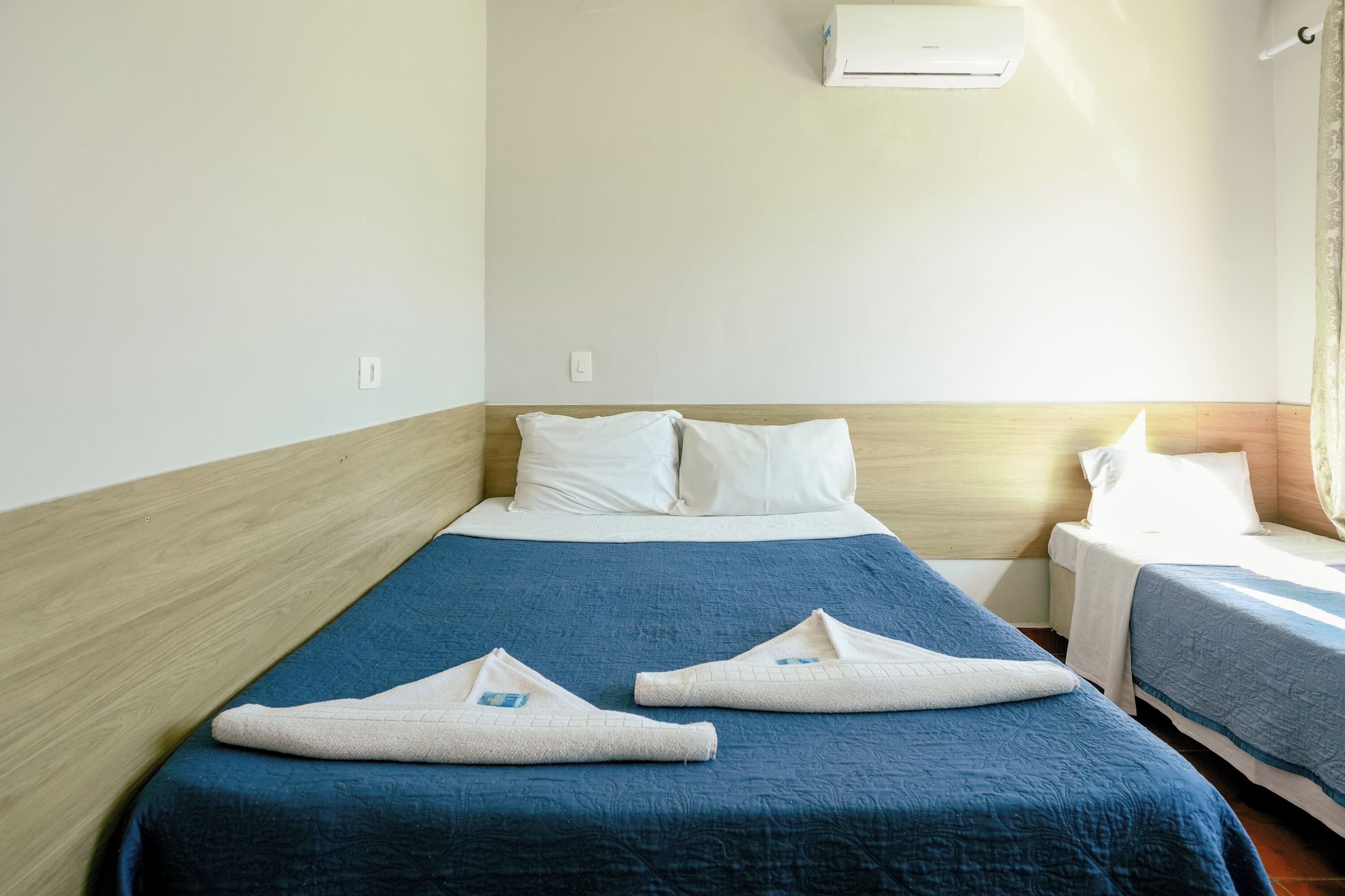 A hotel room with a large bed, two smaller beds, a wooden headboard, white pillows, a blue bedspread, and a white air conditioning unit.