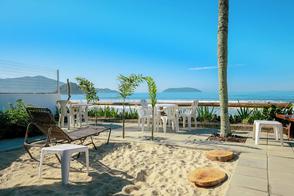 Une terrasse au bord de la plage, avec des chaises en plastique blanc, un fauteuil en osier et une petite table.