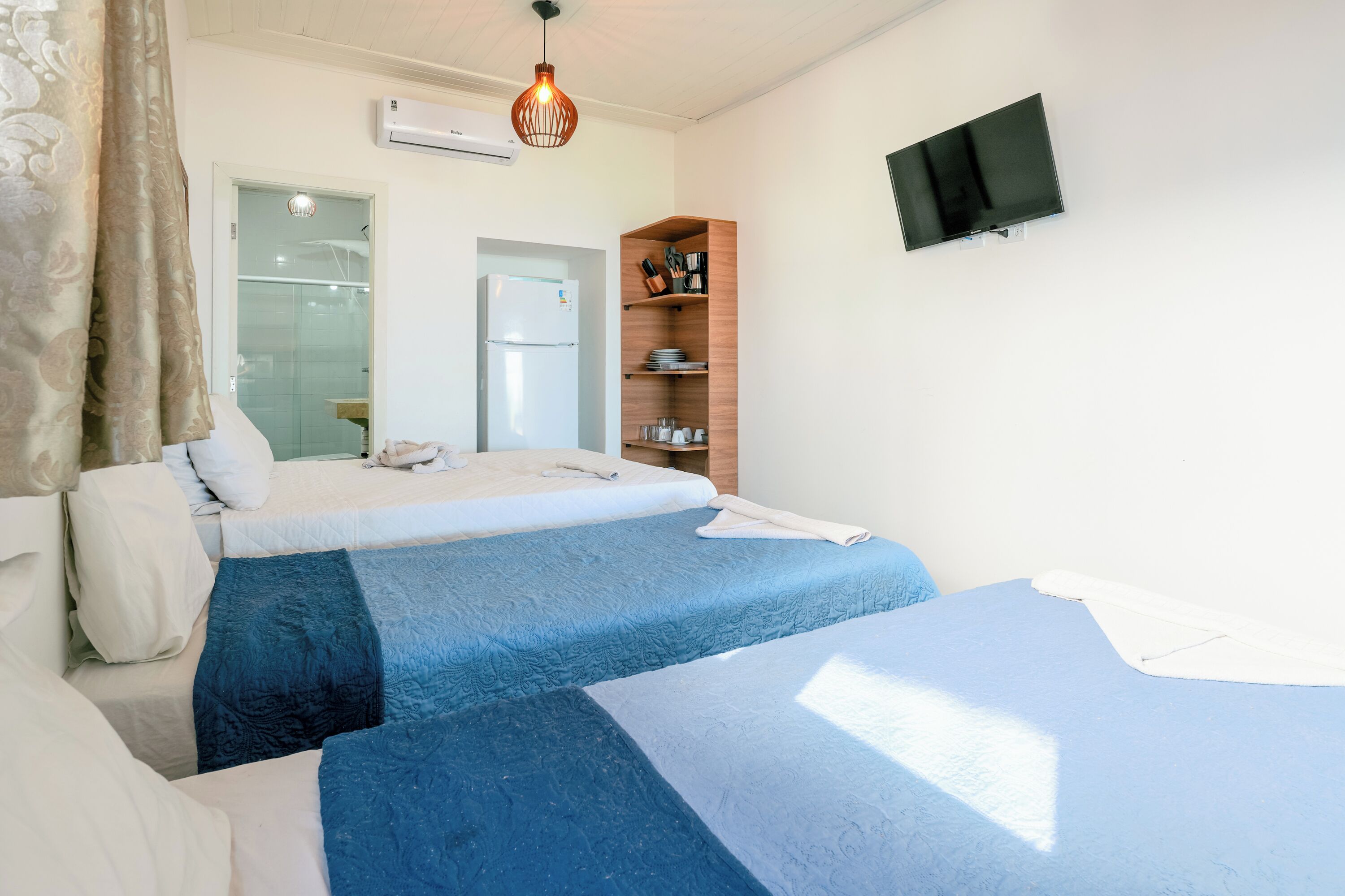 A hotel room with two beds, a TV mounted on the wall, a wooden shelf with kitchenware, and a door leading to a bathroom.