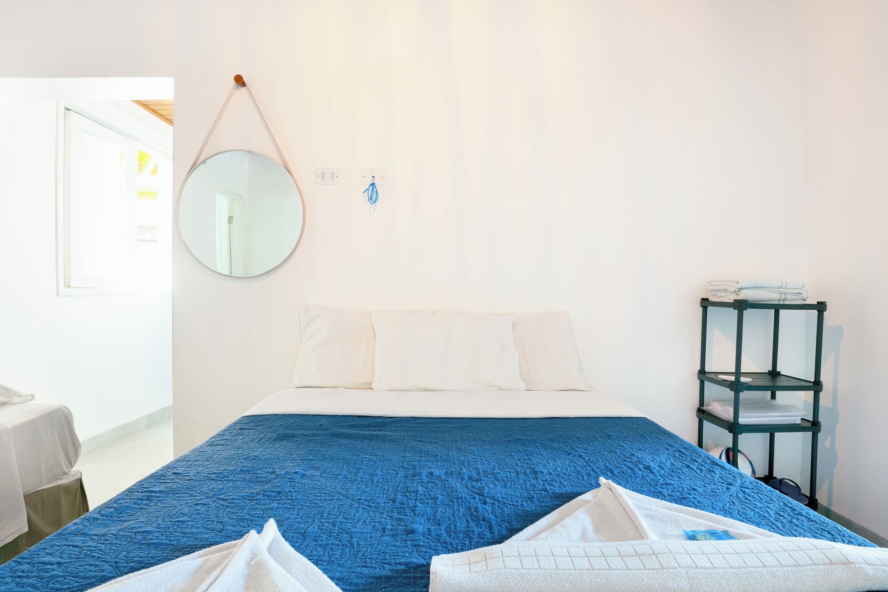 Une chambre d’hôtel avec un lit, une couvre-lit bleue, des oreillers blancs, un miroir rond et une étagère verte où sont pliées des serviettes.