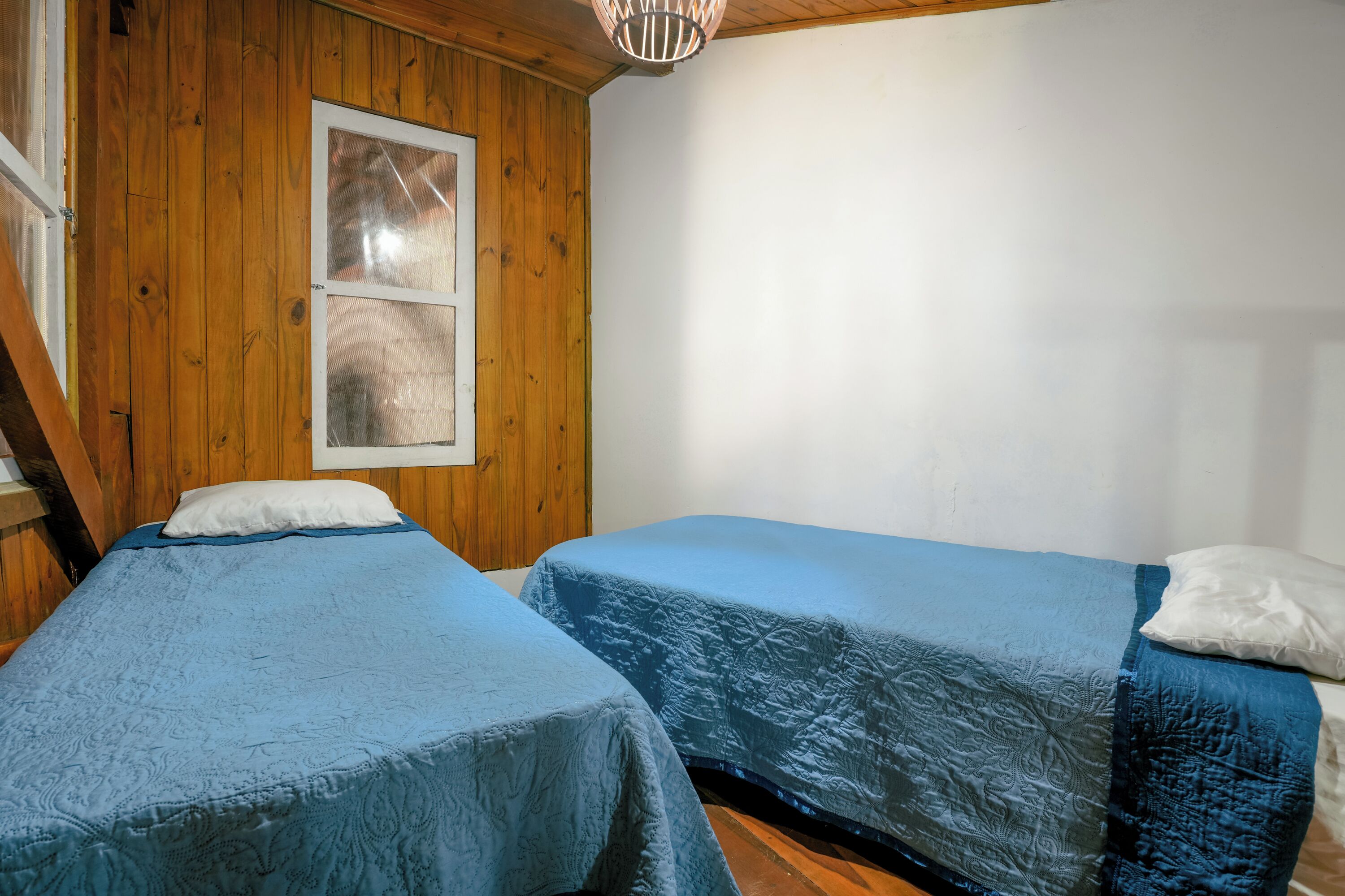 A room with two beds, wooden walls, a window, and a hanging light fixture.