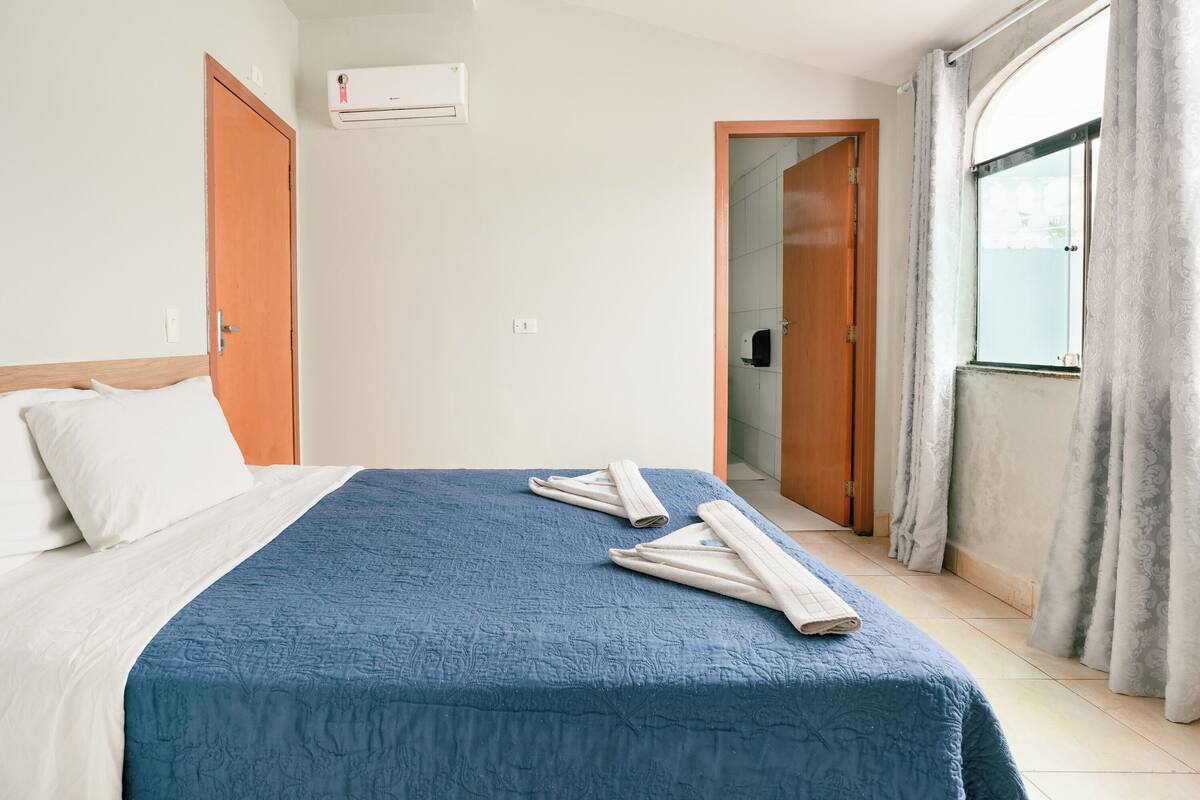 Une chambre d’hôtel avec un lit, une couvre-lit bleue, deux serviettes enroulées, une porte en bois, un appareil de climatisation et une fenêtre avec des rideaux.