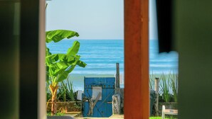 Uitzicht door een deuropening op een strand met een blauw hek, een bankje en een bananenplant.