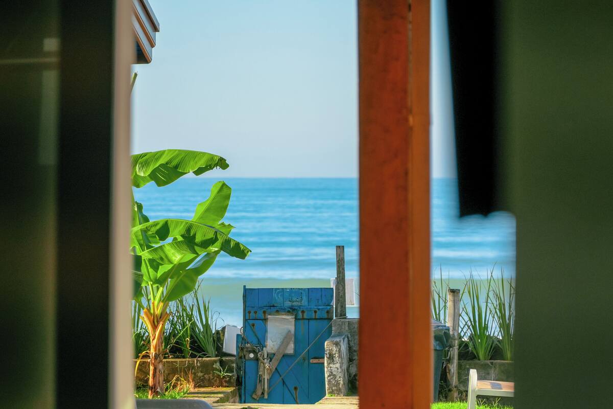 Une vue à travers une porte donnant sur une plage, avec une porte bleue, un banc et un bananier.
