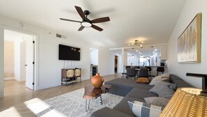 A spacious living area with a ceiling fan, a flat-screen TV mounted on the wall, a dining area with chairs, and a kitchen in the background.