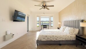 A spacious bedroom with a large bed, a ceiling fan, a flat-screen TV, and a view of the outdoors through large windows.