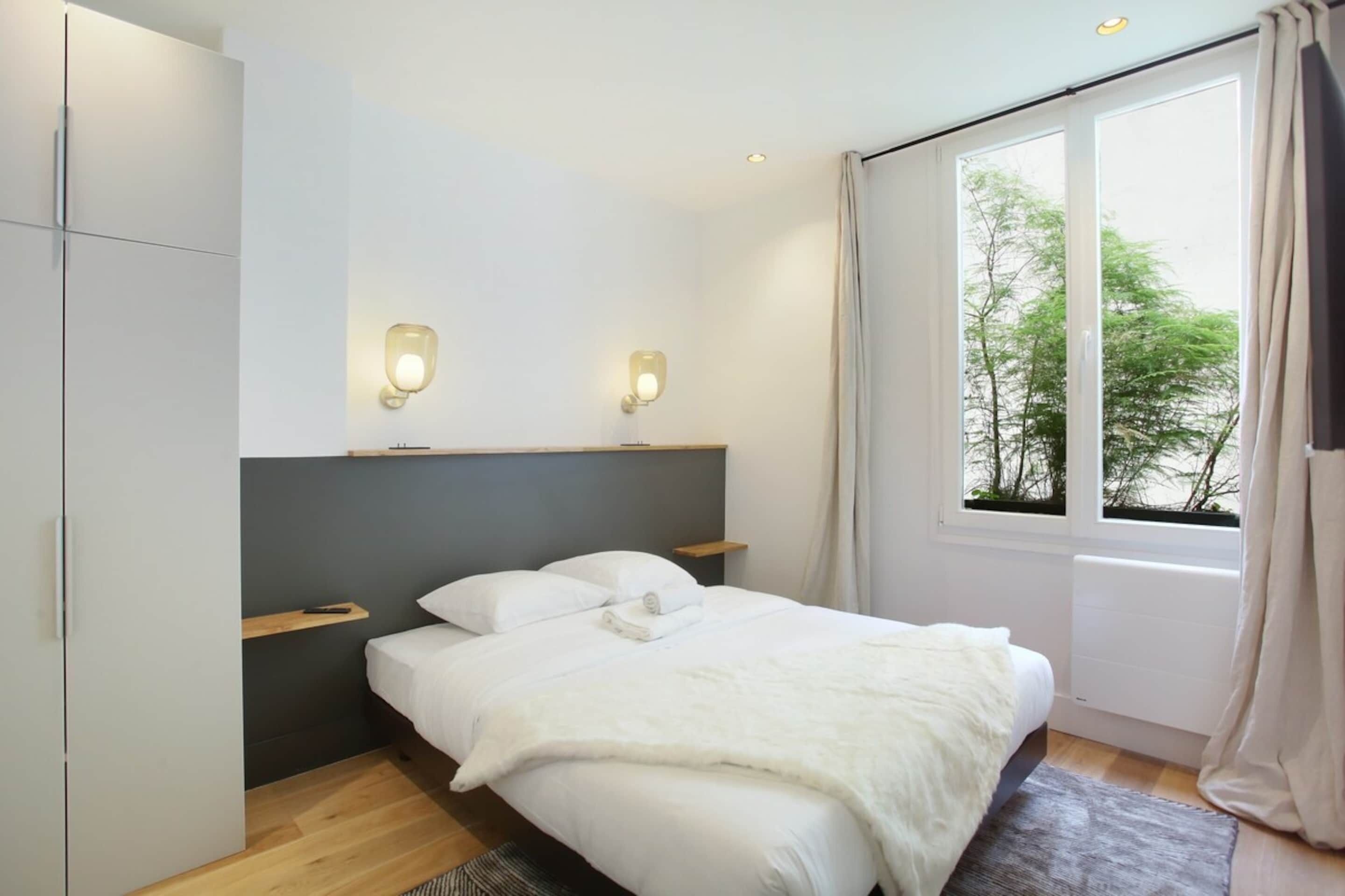 A bedroom with a bed, bedside tables, a wardrobe, a window with curtains, and wall-mounted lights.