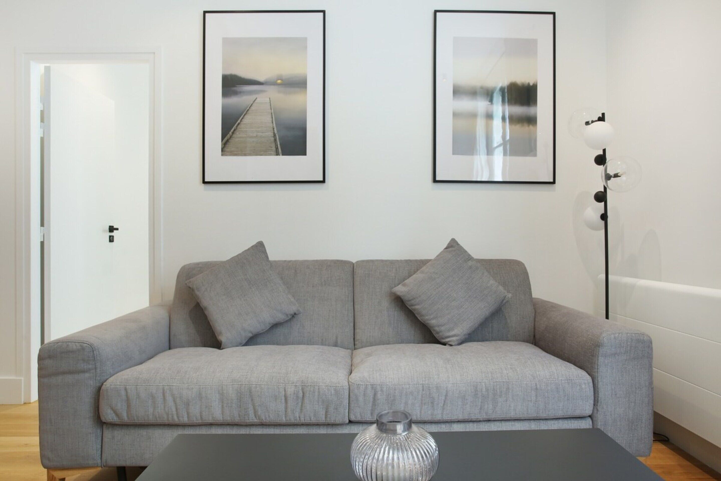 A modern living room with a grey sofa, two framed pictures on the wall, a floor lamp, and a vase on a black table.