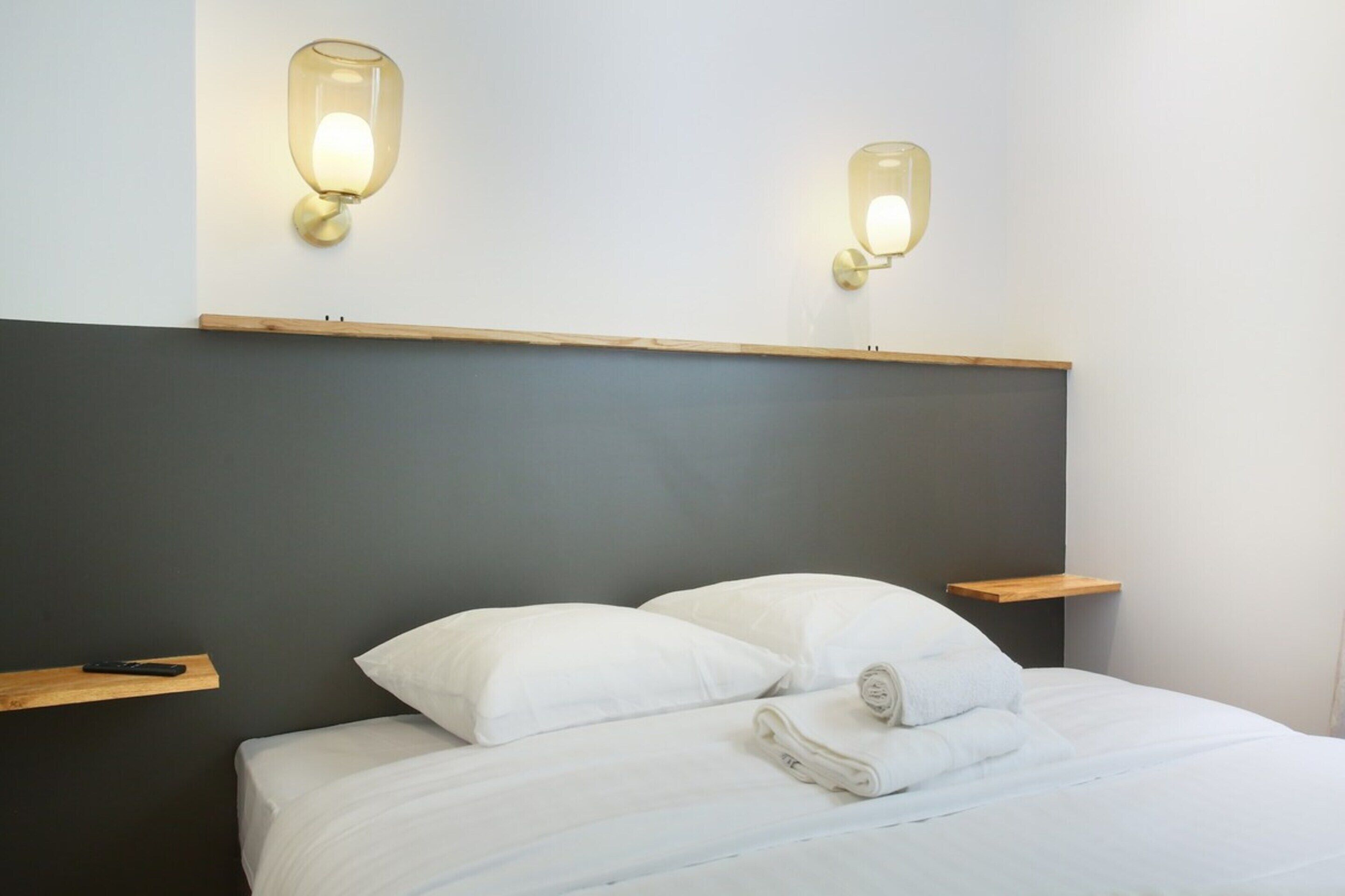 A neatly made bed with white linens and pillows, two bedside tables with a remote control on one, and wall-mounted light fixtures above the bed.