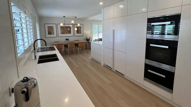 Een moderne keuken met witte kasten, een inbouwoven en een eethoek met een houten tafel en stoelen.