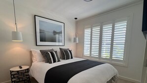 Een slaapkamer met een bed, twee bedlampjes, een ingelijst beeld aan de muur en een raam met jaloezieën.