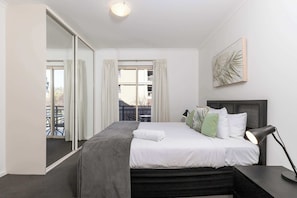 A bedroom with a bed, a bedside table, a lamp, a large mirror, and a view of the outside. - Park Views on Hurtle: CBD, balcony, tennis court (Adelaide)