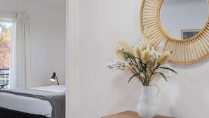 Ett sovrum med en säng, en sänglampa, ett fönster med gardiner, en spegel och en vas med blommor på ett träbord bredvid sängen.