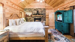 A wooden cabin bedroom with a stone fireplace, a bed with white bedding, a ceiling fan, and a teal cabinet. - Leaping Lizard Lodge | Sleeps 16 | 4 Beds | 3.5 Baths (Broken Bow)