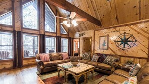 A cozy cabin interior with a wooden ceiling, large windows, a leather sofa set, a coffee table, and a compass wall decoration. - Leaping Lizard Lodge | Sleeps 16 | 4 Beds | 3.5 Baths (Broken Bow)