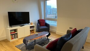 A modern living room with a flat-screen TV, a bookshelf, a grey sofa with red and blue cushions, a coffee table, and a large window with a view of a snowy landscape. - 2br-modern-fullkitchen-25mincentrallondon-parking (Rickmansworth)