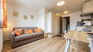 Een moderne woonkamer met een grijze bank, een klein witte tafeltje en een houten eettafel. Er is een keukenhoek met een koffieapparaat en een waterkoker.