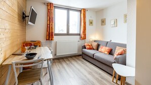 Een knus vertrek met een houten wand, een grijze bank, een eettafel met stoelen en een raam met gordijnen.