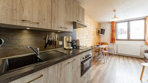 Een moderne keuken met houten kastjes, een inbouwoven en een koffieapparaat. Er is een eethoek met een tafel en stoelen, en een raam met gordijnen.