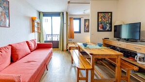 A cozy living room with a red sofa, wooden dining table, and benches. A television is on a wooden cabinet, and there are paintings on the walls.