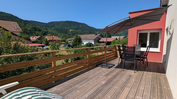 Una terraza de madera con una mesa y sillas, una valla de madera, y vistas a casas y montañas al fondo.