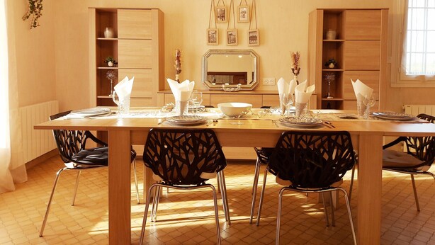 Un comedor con una larga mesa de madera dispuesta para cuatro personas, rodeada de sillas negras con un diseño geométrico. Hay armarios de madera a cada lado de la mesa, un espejo encima y una guirnalda de luces colgando del techo.