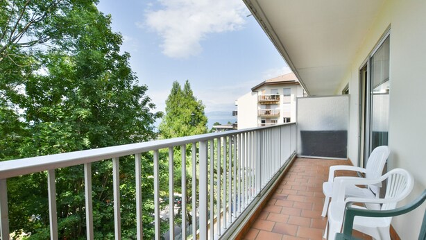 Un balcon avec des chaises blanches, un sol carrelé et une vue sur les arbres et les bâtiments.