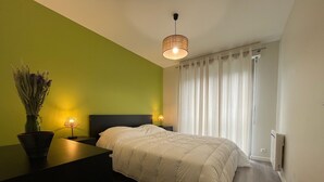 Une chambre à coucher avec un lit, une table de chevet, une lampe, un vase contenant des fleurs séchées et une fenêtre avec des rideaux.
