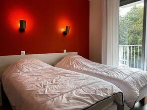 Ein Zimmer mit roter Wand, zwei Betten mit weißer Bettwäsche und einem Fenster mit Blick auf Bäume.