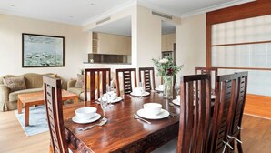 A dining area with a wooden table set for six, surrounded by chairs, with a kitchen in the background.
