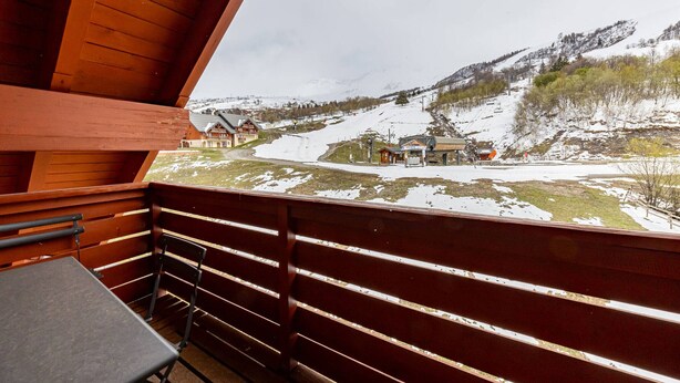 Un balcón de madera con mesa y sillas, con vistas a un paisaje montañoso nevado con edificios y un telesilla.