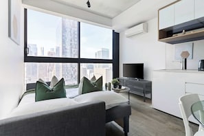 A modern apartment with a large window offering a city view, a kitchenette with a TV, and a seating area with cushions.