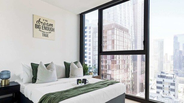 A modern bedroom with a large window, a bed with green and white bedding, a bedside table with a lamp, and a motivational quote on the wall.