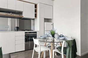 A modern kitchen with white cabinetry, a glass dining table, and a green plant. - Pet-friendly Stylish 2BR Apt, Panoramic City Views (Melbourne)