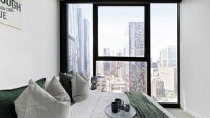 A high-rise room with a city view, a bed with green cushions, a tray with two mugs, and a small plant.