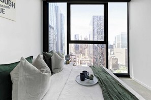 Une chambre située dans un immeuble de grande hauteur, avec vue sur la ville, un lit agrémenté de coussins verts, un plateau avec deux tasses et une petite plante.