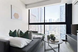 A modern living room with a large window, a grey sofa, and a small round table with a tray and potted plants.