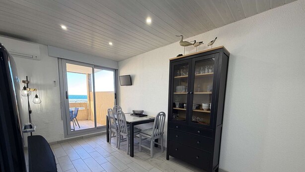 Un coin repas avec une table et des chaises, un meuble avec des portes en verre, et une vue sur un balcon extérieur accessible par une porte coulissante.