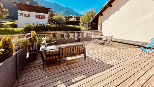 Eine Holzterrasse mit Gartenmöbeln, ein weißes Gebäude und Blick auf die Berge.