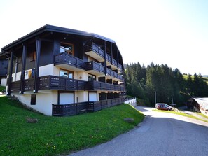 Un edificio de varios pisos con balcones, un camino pavimentado y un paisaje boscoso al fondo.