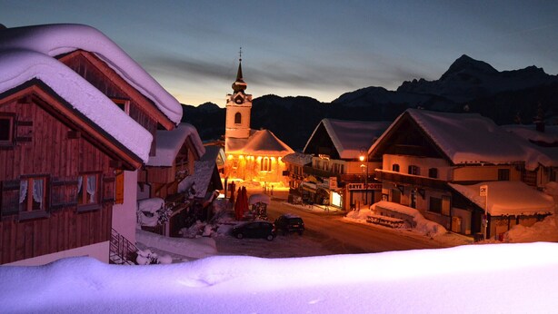 Un village enneigé, avec ses bâtiments traditionnels en bois, une église dotée d’un clocher et une route qui traverse le village.