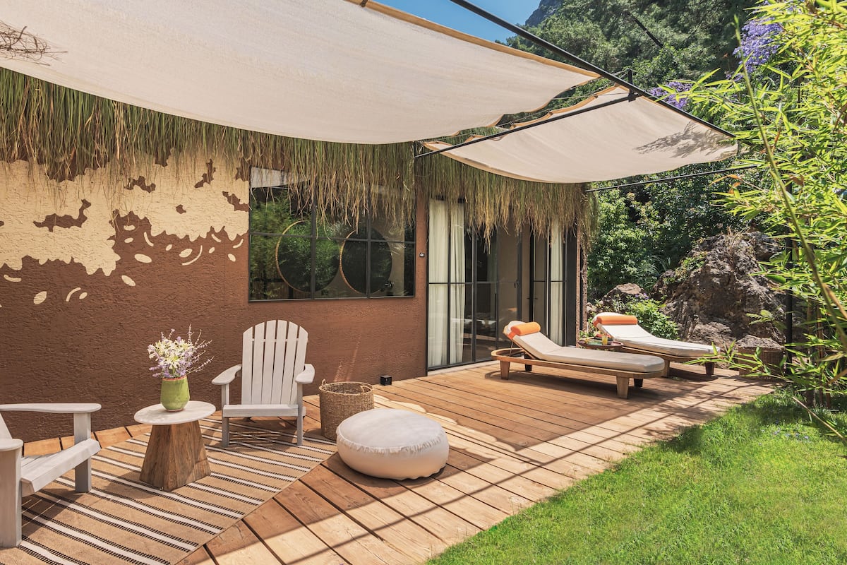 A patio with two Adirondack chairs, a round ottoman, a small table with a vase of flowers, and a large umbrella providing shade.