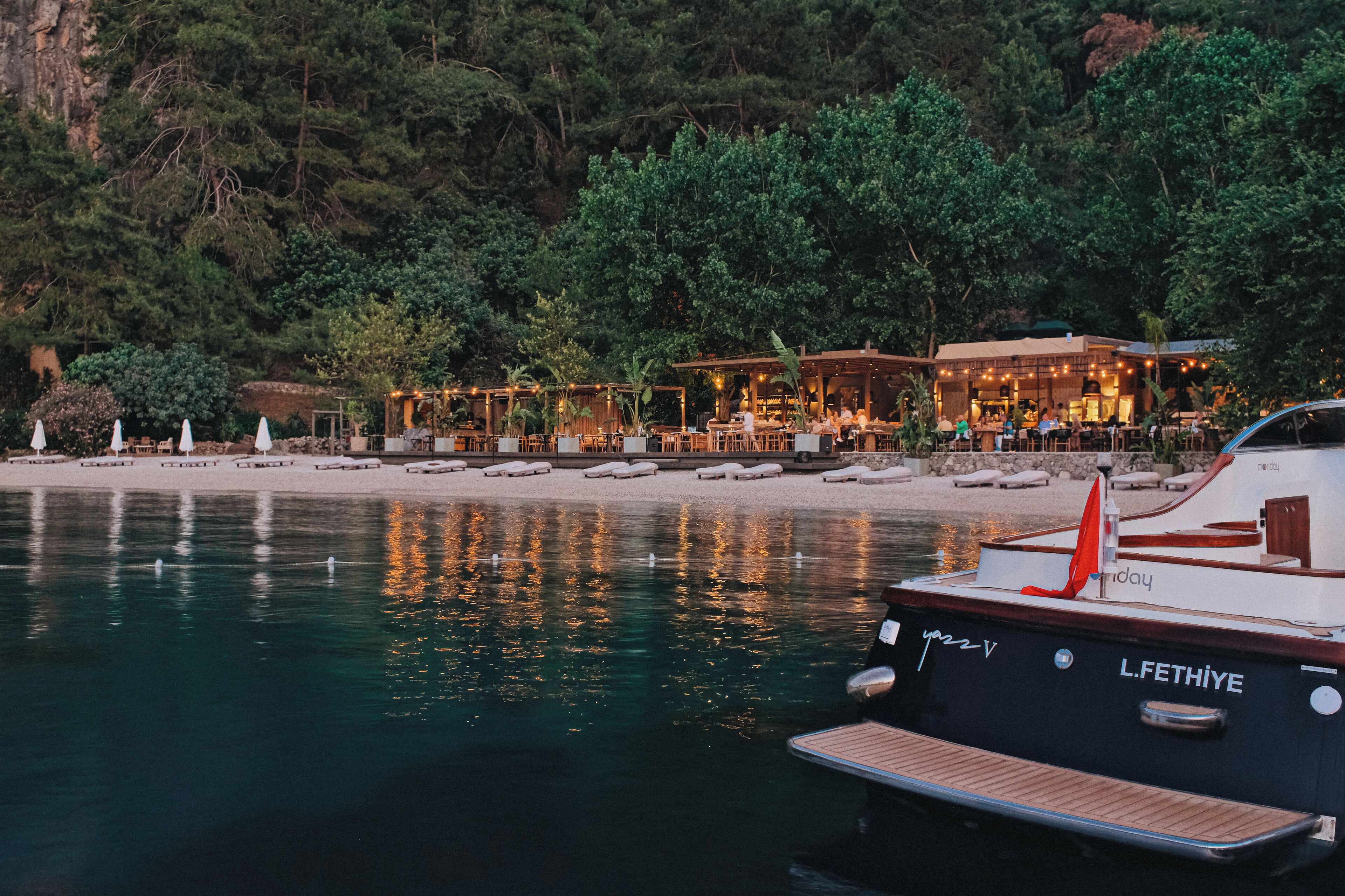 A waterfront restaurant with outdoor seating, a boat docked nearby, and a forested hillside in the background.