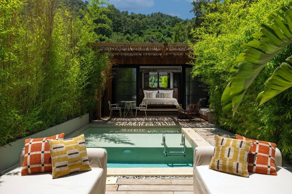 A pool area with a bed and seating area, surrounded by lush greenery.